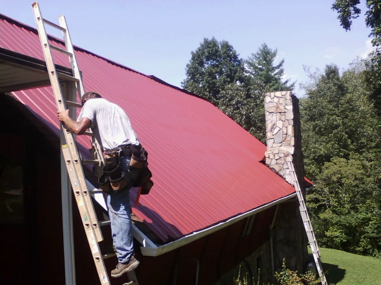 Roofer on a ladder beside a red metal roof and stone chimney — Soffit & Fascia Repair by Roofing Brothers in Bouckville