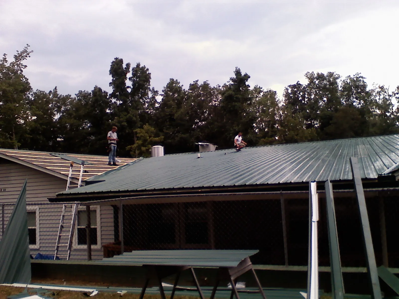 Two roofers installing green metal panels above a covered porch in Bouckville